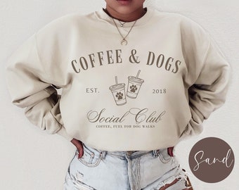 Puede incluir: Una sudadera beige con el texto "COFFEE & DOGS EST. 2018 Social Club COFFEE, FUEL FOR DOG WALKS" impreso en la parte delantera. La sudadera presenta un gráfico de dos tazas de café con huellas de patas.