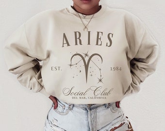 Op de afbeelding: Een beige sweatshirt met de tekst "ARIES" in bruine letters, een gestileerd sterrenbeeld symbool en de tekst "Social Club Del Mar, California" in bruin cursief schrift. Het sweatshirt heeft ook de tekst "EST. 1984" in bruine letters.