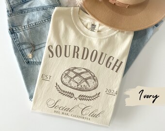May include: Ivory t-shirt with a brown graphic of a sourdough loaf of bread and the text "SOURDOUGH Social Club EST 2024 DEL MAR, CALIFORNIA".
