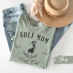 K&ouml;nnte beinhalten: Ein hellgr&uuml;nes T-Shirt mit dem Text "GOLF MOM EST. 2024 Social Club TORREY PINES" und einer Schwarzwei&szlig;illustration einer Frau, die Golf spielt.