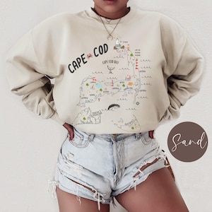 May include: A beige sweatshirt with a graphic print of a map of Cape Cod, Massachusetts. The map includes various towns and landmarks, including Provincetown, Chatham, and Martha's Vineyard. The text "CAPE COD" is printed in black at the top of the map.