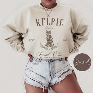 May include: A beige sweatshirt with the text "KELPIE Bailey EST. 2024 Social Club DEL MAR, CALIFORNIA" and a graphic of a dog. The sweatshirt is being worn by a person.