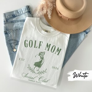 K&ouml;nnte beinhalten: Wei&szlig;es T-Shirt mit gr&uuml;nem Text, der "GOLF MOM EST. 2024 Social Club TORREY PINES" lautet. Eine gr&uuml;ne Illustration einer Frau, die Golf spielt, ist mittig auf dem T-Shirt platziert.