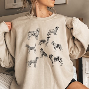 K&ouml;nnte beinhalten: Ein beiger Pullover mit einem grafischen Druck verschiedener Hunderassen in Schwarzwei&szlig;. Der Pullover hat einen Rundhalsausschnitt und lange &Auml;rmel.