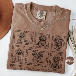 May include: Espresso-colored t-shirt featuring a postage stamp design with illustrations of puppies. The shirt has a crew neck and short sleeves. A silver necklace with a small pendant is draped on the shirt.