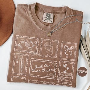 May include: Espresso-colored t-shirt with a white graphic of postage stamp designs, including a coffee cup, bow, and books. The text "Just One More Chapter" is featured. A silver necklace and a brown hat are also visible.