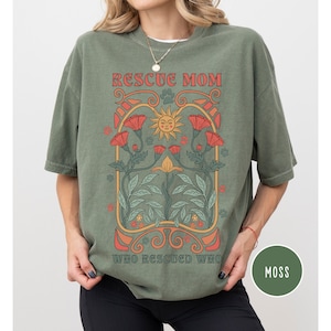 May include: A green t-shirt with a graphic design featuring a sun, flowers, and the text "Rescue Mom Who Rescued Who". The shirt is a relaxed fit and has a distressed look.