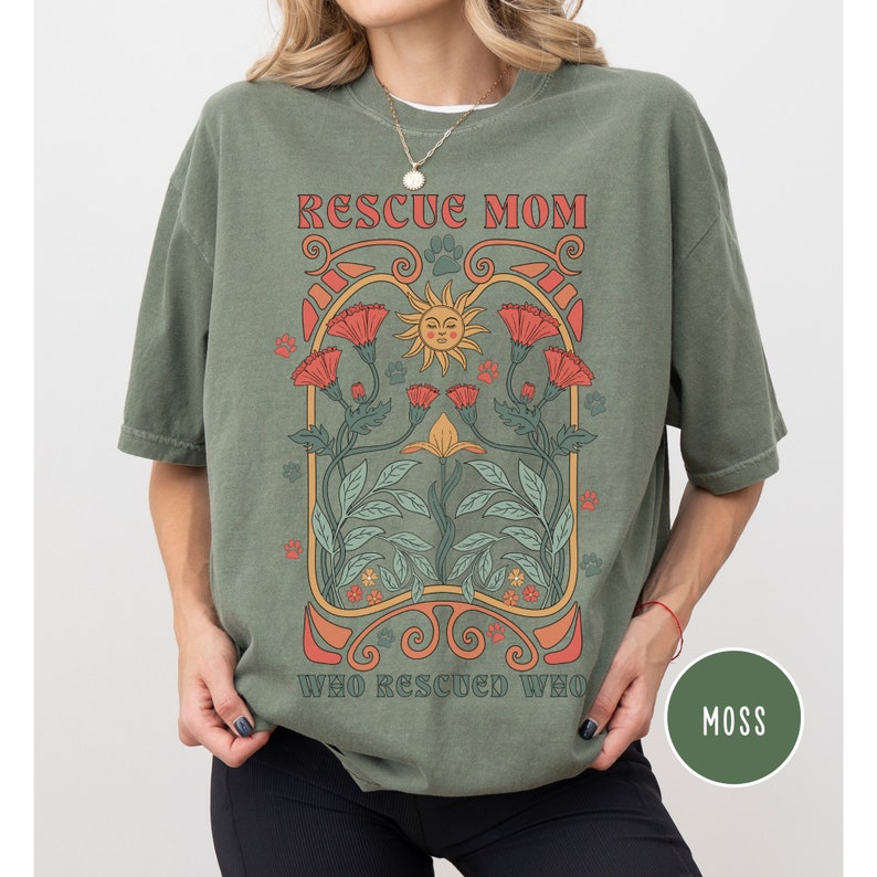 May include: A green t-shirt with a graphic design featuring a sun, flowers, and the text "Rescue Mom Who Rescued Who". The shirt is a relaxed fit and has a distressed look.