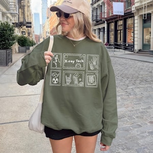 May include: An olive green sweatshirt with a white graphic design featuring the text "X-ray Tech" and various medical-themed illustrations. The sweatshirt has long sleeves and a crew neck.