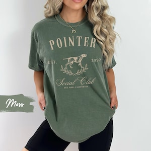 May include: A green t-shirt with a vintage-style graphic design featuring a dog, the text "POINTER EST. 1992 Social Club DEL MAR, CALIFORNIA", and a laurel wreath.