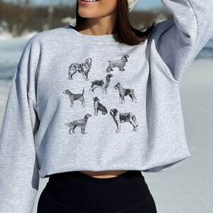 K&ouml;nnte beinhalten: Ein grauer, kurzer Sweatshirt mit einem Aufdruck verschiedener Hunderassen in Schwarzwei&szlig;. Der Sweatshirt hat einen Rundhalsausschnitt und lange &Auml;rmel.