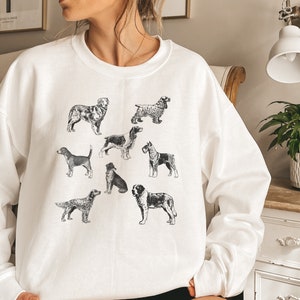 K&ouml;nnte beinhalten: Wei&szlig;es Sweatshirt mit einer Schwarzwei&szlig;illustration verschiedener Hunderassen. Die Hunde sind in verschiedenen Posen und Gr&ouml;&szlig;en dargestellt und alle nach vorne gerichtet.