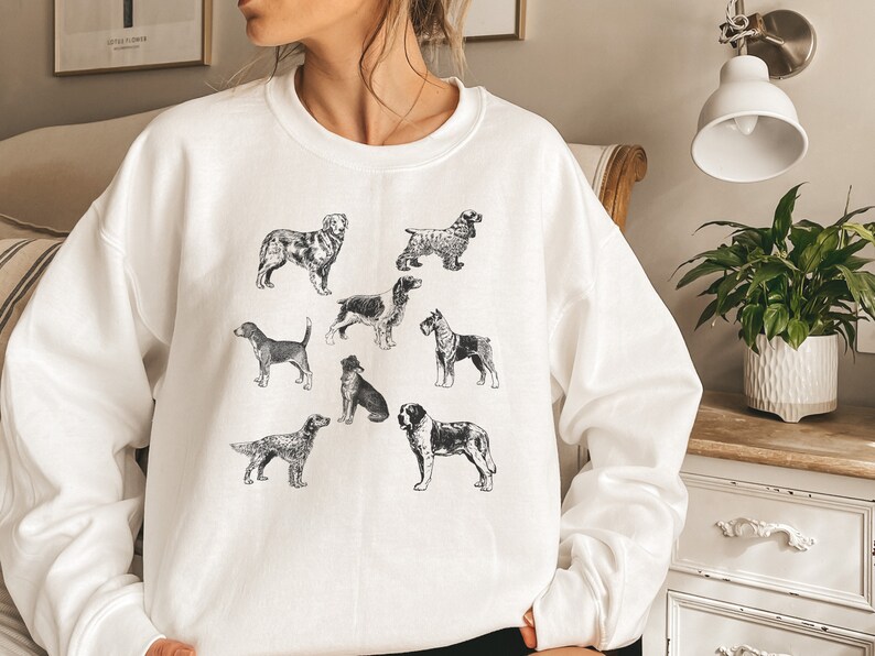 K&ouml;nnte beinhalten: Wei&szlig;es Sweatshirt mit einer Schwarzwei&szlig;illustration verschiedener Hunderassen. Die Hunde sind in verschiedenen Posen und Gr&ouml;&szlig;en dargestellt und alle nach vorne gerichtet.