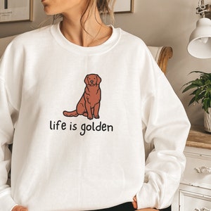 Op de afbeelding: Witte trui met een illustratie van een bruine golden retriever en de tekst "life is golden" gedrukt op de voorkant.