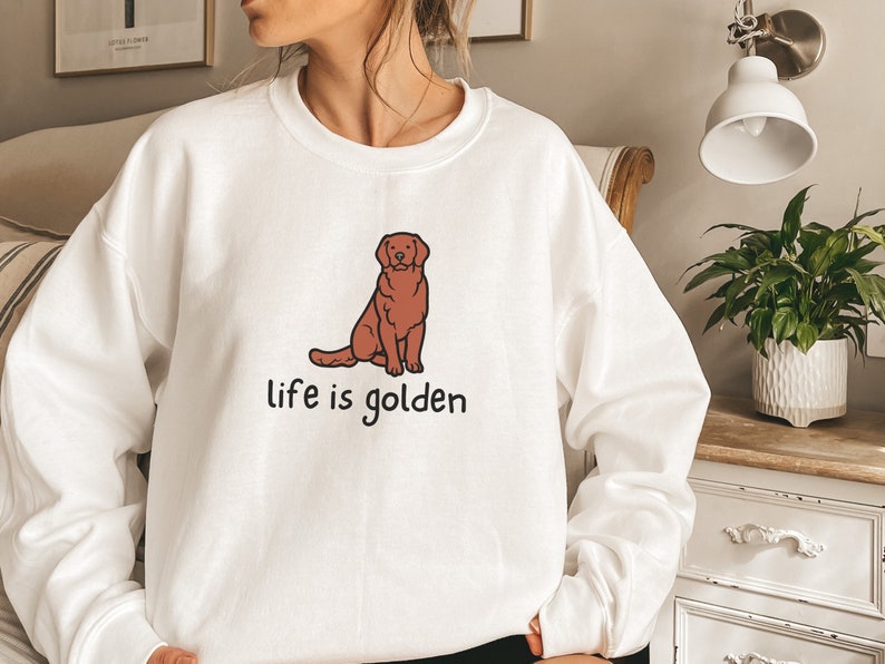 Op de afbeelding: Witte trui met een illustratie van een bruine golden retriever en de tekst "life is golden" gedrukt op de voorkant.