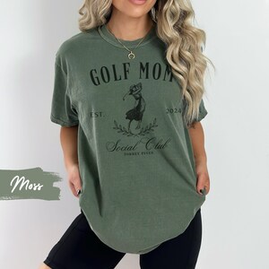 K&ouml;nnte beinhalten: Ein gr&uuml;nes T-Shirt mit dem Text "GOLF MOM EST. 2024 Social Club TORREY PINES" und einer Grafik einer Frau, die Golf spielt.
