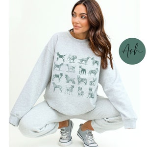 May include: A woman wearing a grey sweatshirt with a graphic print of various dog breeds. The sweatshirt has a crew neck and long sleeves. The woman is wearing grey sweatpants and white sneakers.