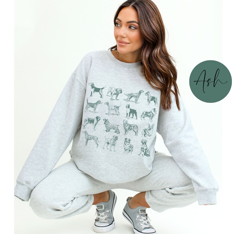 May include: A woman wearing a grey sweatshirt with a graphic print of various dog breeds. The sweatshirt has a crew neck and long sleeves. The woman is wearing grey sweatpants and white sneakers.