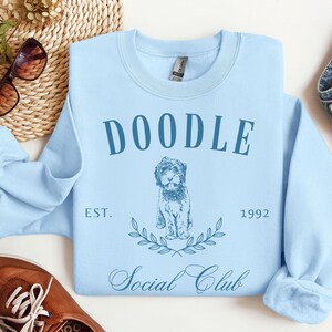Puede incluir: Sudadera azul claro con el texto "Doodle Social Club Est. 1992" y una ilustraci&oacute;n de un perro doodle.