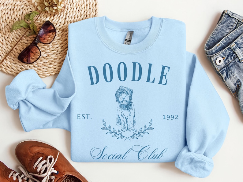 Puede incluir: Sudadera azul claro con el texto "Doodle Social Club Est. 1992" y una ilustraci&oacute;n de un perro doodle.