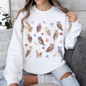 May include: White sweatshirt with a colorful print of owls, butterflies, and flowers. The owls are in various poses and have different patterns.