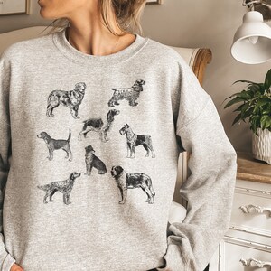 K&ouml;nnte beinhalten: Ein graues Sweatshirt mit einer Schwarzwei&szlig;illustration verschiedener Hunderassen. Die Hunde sind in einem Rastermuster auf der Vorderseite des Sweatshirts angeordnet.