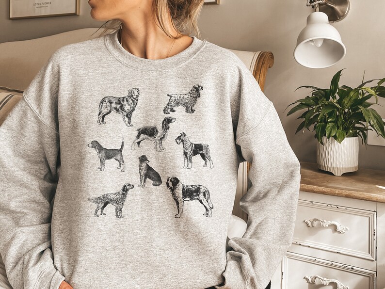 K&ouml;nnte beinhalten: Ein graues Sweatshirt mit einer Schwarzwei&szlig;illustration verschiedener Hunderassen. Die Hunde sind in einem Rastermuster auf der Vorderseite des Sweatshirts angeordnet.