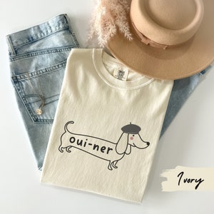 May include: A white t-shirt with a black and white illustration of a dachshund wearing a beret. The dachshund is labeled "oui-ner". The shirt is on a light blue denim background with a brown hat and a white feather boa.