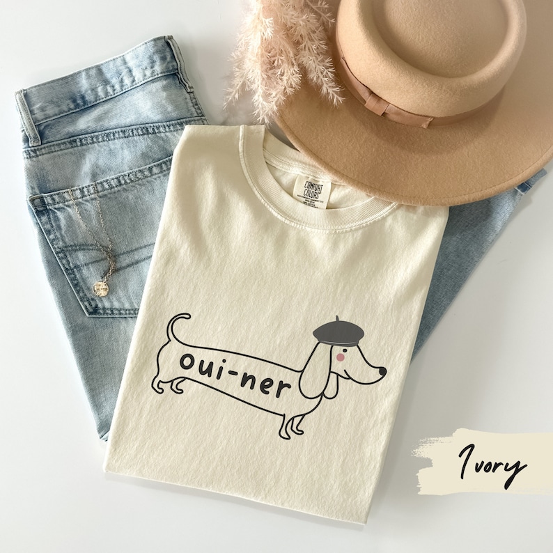 May include: A white t-shirt with a black and white illustration of a dachshund wearing a beret. The dachshund is labeled "oui-ner". The shirt is on a light blue denim background with a brown hat and a white feather boa.