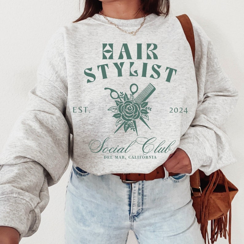 K&ouml;nnte beinhalten: Grauer Pullover mit dem Text "HAIR STYLIST" in Gr&uuml;n, einem floralen Design mit Schere und Kamm und dem Text "Social Club Del Mar, Kalifornien" in Kursivschrift.