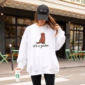 Op de afbeelding: Witte trui met een illustratie van een bruine golden retriever en de tekst "life is golden". Het model draagt een zwarte baseballpet met de tekst "11:11" erop.
