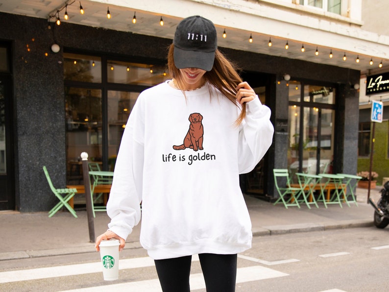 Op de afbeelding: Witte trui met een illustratie van een bruine golden retriever en de tekst "life is golden". Het model draagt een zwarte baseballpet met de tekst "11:11" erop.