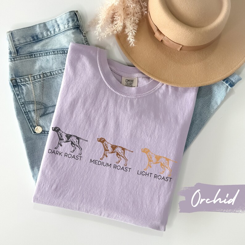 Op de afbeelding: Een lichtpaarse T-shirt met een afbeelding van drie honden die verschillende koffieroasters vertegenwoordigen: donker gebrande koffie, medium gebrande koffie en licht gebrande koffie. De tekst "COMFORT COLORS" is zichtbaar op het label van het shirt. Het shirt ligt op een wit oppervlak met een blauwe spijkerbroek en een bruine hoed.