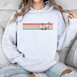May include: Light gray hoodie with a graphic of two Siamese cats on a horizontal striped design in red, orange, tan, and teal. The hoodie has a drawstring hood and long sleeves.