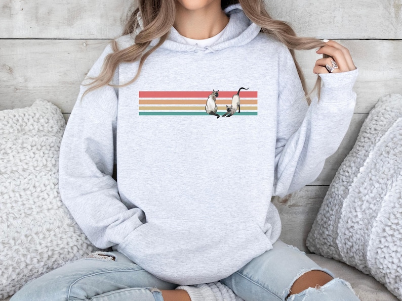 May include: Light gray hoodie with a graphic of two Siamese cats on a horizontal striped design in red, orange, tan, and teal. The hoodie has a drawstring hood and long sleeves.