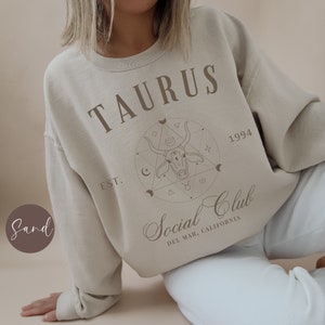 Puede incluir: Una sudadera beige con el texto "TAURUS EST. 1994 Social Club DEL MAR, CALIFORNIA" y un gr&aacute;fico de una cabeza de toro dentro de un c&iacute;rculo con formas geom&eacute;tricas.