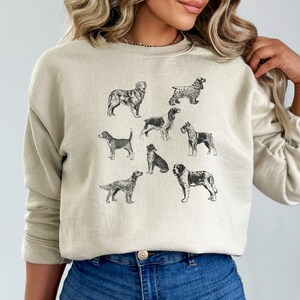 K&ouml;nnte beinhalten: Ein hellbeiger Pullover mit einem grafischen Druck verschiedener Hunderassen in Schwarzwei&szlig;. Die Hunde sind in einem Vintage-Illustrationsstil dargestellt.