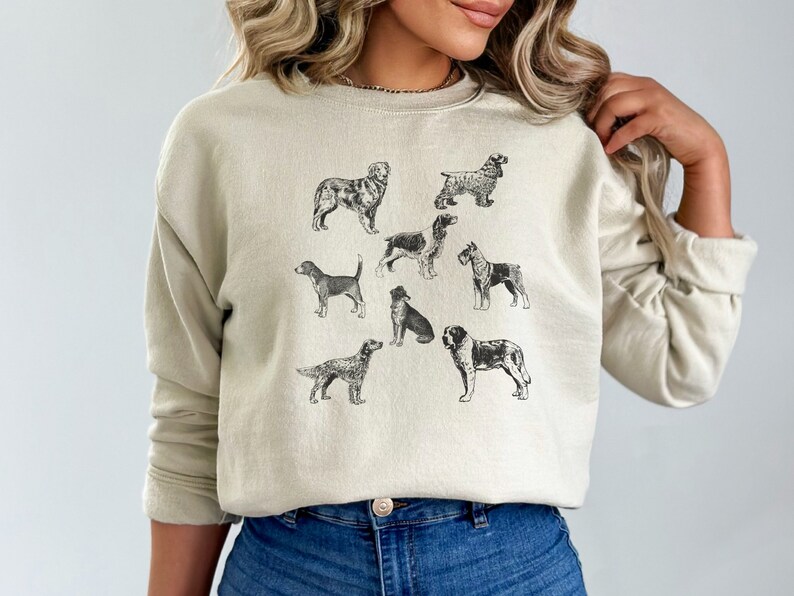 K&ouml;nnte beinhalten: Ein hellbeiger Pullover mit einem grafischen Druck verschiedener Hunderassen in Schwarzwei&szlig;. Die Hunde sind in einem Vintage-Illustrationsstil dargestellt.