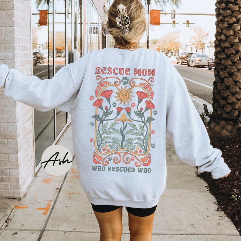 Puede incluir: Sudadera gris con un dise&ntilde;o floral y el texto "Rescue Mom Who Rescued Who" impreso en la espalda.