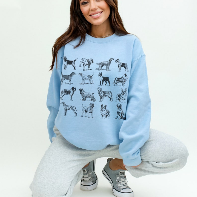 May include: A light blue sweatshirt with a graphic print of various dog breeds in black and white. The sweatshirt is worn by a person with long brown hair and is styled with grey sweatpants.