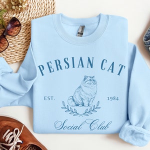 May include: Light blue sweatshirt featuring a graphic of a Persian cat and the text "Persian Cat Social Club Est. 1984".