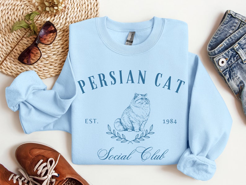 May include: Light blue sweatshirt featuring a graphic of a Persian cat and the text "Persian Cat Social Club Est. 1984".