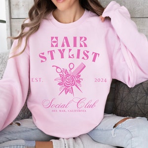 K&ouml;nnte beinhalten: Rosafarbener Pullover mit dem Text "Hair Stylist" und einer Grafik von einer Schere und einem Kamm, die &uuml;ber einer Rose gekreuzt sind. Der Text "Est. 2024" und "Social Club Del Mar, Kalifornien" ist ebenfalls enthalten.