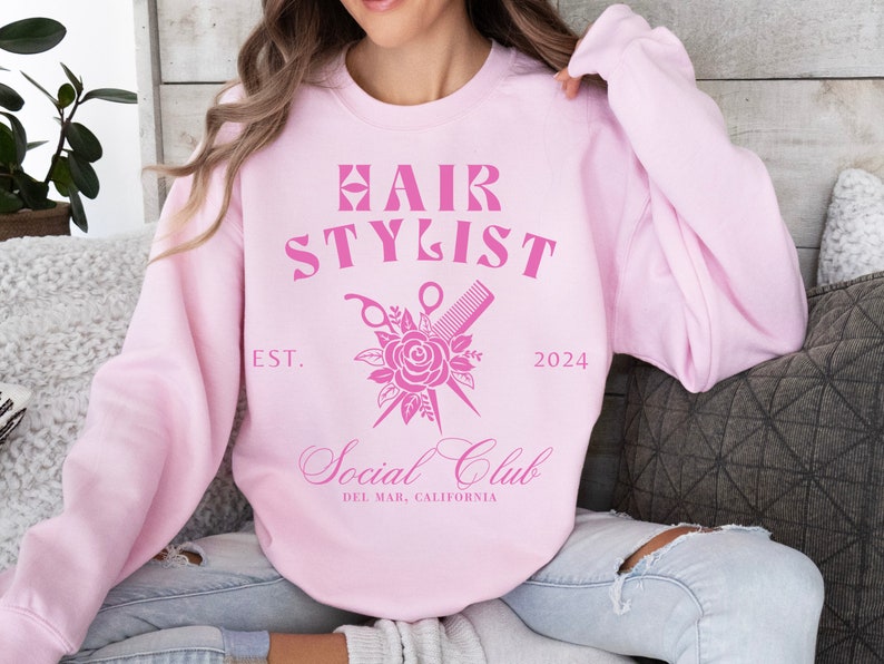 K&ouml;nnte beinhalten: Rosafarbener Pullover mit dem Text "Hair Stylist" und einer Grafik von einer Schere und einem Kamm, die &uuml;ber einer Rose gekreuzt sind. Der Text "Est. 2024" und "Social Club Del Mar, Kalifornien" ist ebenfalls enthalten.