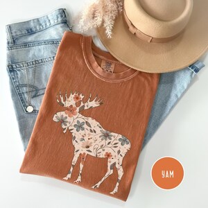 May include: A rust-coloured t-shirt with a floral moose design, alongside light-wash jeans and a tan hat. A gold necklace and dried flowers are also visible. The shirt has a round neck and short sleeves. A round orange sticker with the letters "YAM" is in the bottom right corner.