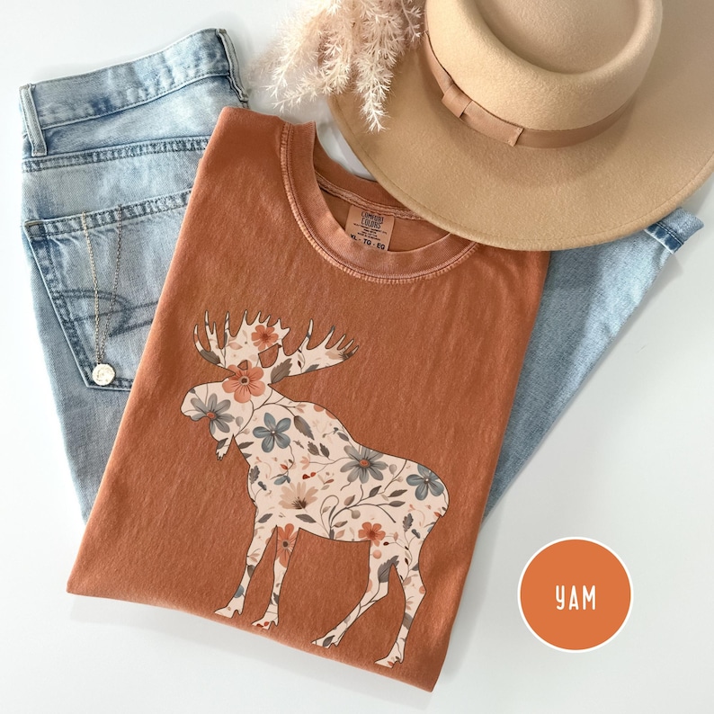 May include: A rust-coloured t-shirt with a floral moose design, alongside light-wash jeans and a tan hat. A gold necklace and dried flowers are also visible. The shirt has a round neck and short sleeves. A round orange sticker with the letters "YAM" is in the bottom right corner.