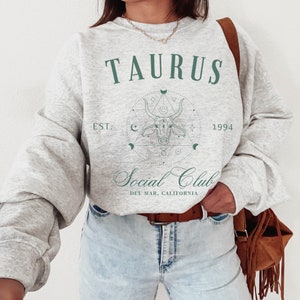 Puede incluir: Sudadera gris con un dise&ntilde;o gr&aacute;fico verde que presenta la palabra "TAURUS" y una cabeza de toro dentro de un c&iacute;rculo con formas geom&eacute;tricas. El texto "EST. 1994" y "Social Club DEL MAR, CALIFORNIA" tambi&eacute;n est&aacute;n incluidos en el dise&ntilde;o.
