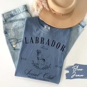 May include: Blue jean t-shirt with a black graphic of a Labrador Retriever, the text "Labrador", "Roxy", "Est. 2024", and "Social Club Del Mar, California".