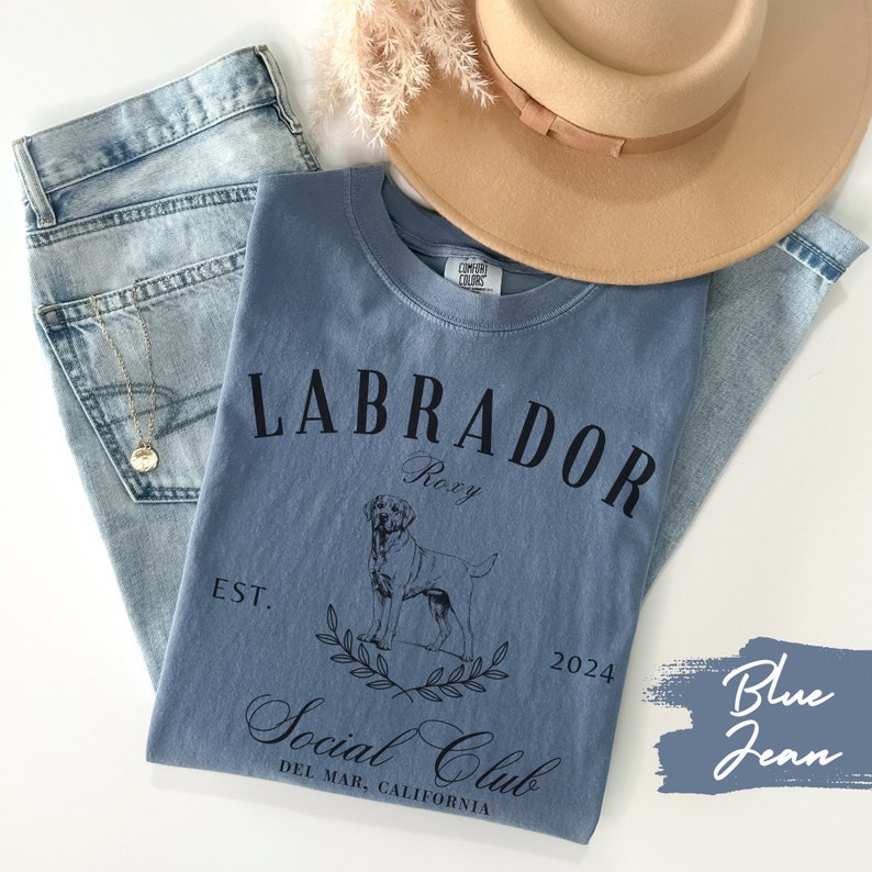 May include: Blue jean t-shirt with a black graphic of a Labrador Retriever, the text "Labrador", "Roxy", "Est. 2024", and "Social Club Del Mar, California".