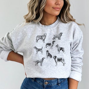 K&ouml;nnte beinhalten: Ein graues Sweatshirt mit einem Aufdruck verschiedener Hunderassen in Schwarzwei&szlig;. Die Hunde sind in einem Kreis auf der Vorderseite des Sweatshirts angeordnet.